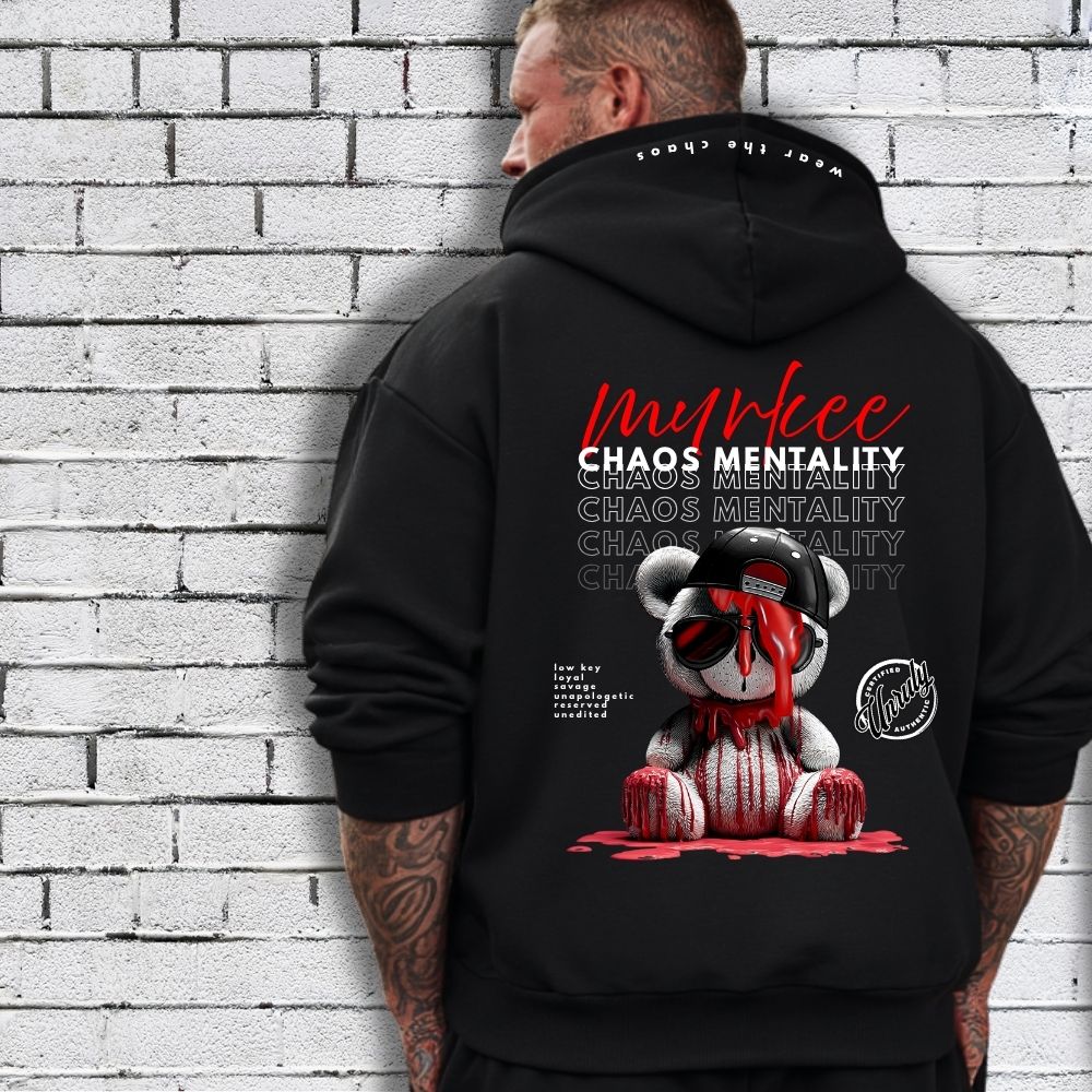 Person wearing a black hoodie with graphic design and text against a white brick wall.