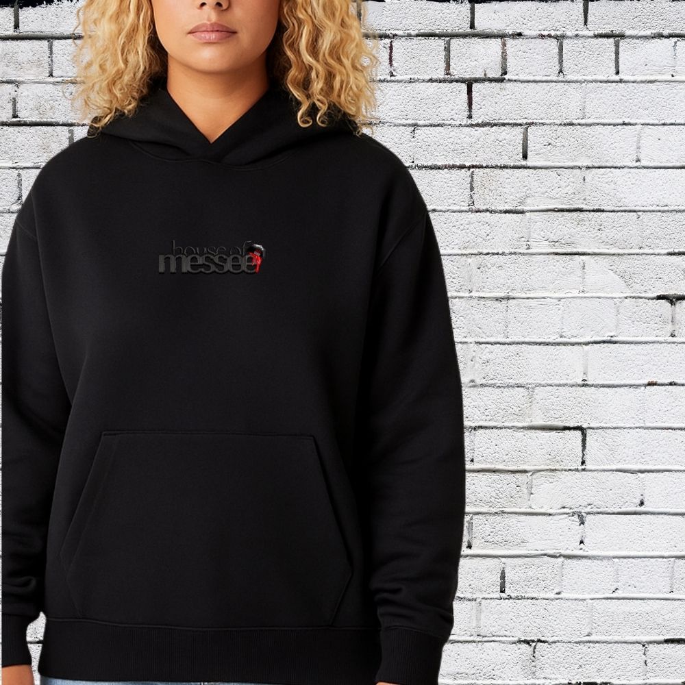 Person wearing a black hoodie with a logo against a white brick wall